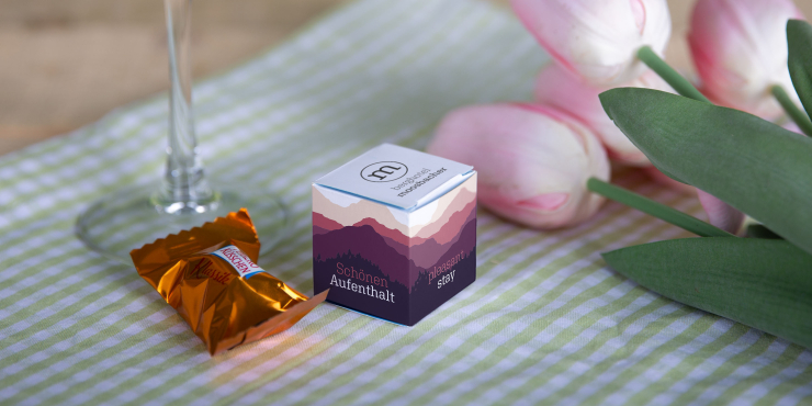 Auf einem gedeckten Tisch steht ein bedruckter Mini-Kartonwürfel, daneben liegt ein Ferrero Küsschen und ein Blumenstrauß mit Tulpen
