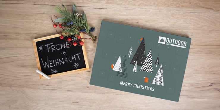 Bedruckter Adventkalender gefüllt mit Schokolade liegt auf einem Tisch neben einer Tafel auf der mit Kreide "Frohe Weihnacht" geschrieben steht
