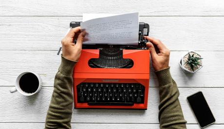 Eine rote Schreibmaschine in welche zwei Hände gerade Papier einspannen.