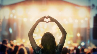 Eine Frau, die mit ihren Händen ein Herz mit Armen über ihrem kopf formt und dabei vor einer Bühne steht.