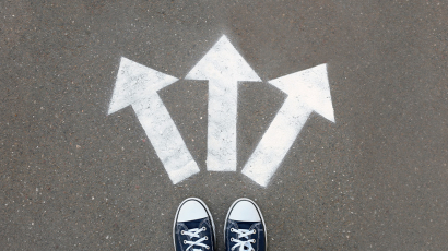 Zwei schwarze Schuhe vor denen drei weiße Pfeile mit Kreide auf den Boden gemalt sind. Diese zeigen von den Schuhen weg in verschiedene Richtungen