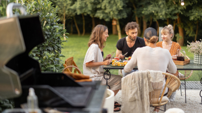 Im Vordergrund steht ein Griller. Im Hintergrund sieht man Menschen zusammen an einem Tisch sitzen.
