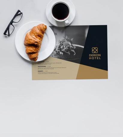 Tischset bedruckt mit Ihrem Logo, welches auf einem grauen Tisch liegt. Darauf steht ein weißer Teller mit einem Croissant darauf und einer Tasse Kaffee und Lesebrille daneben 
