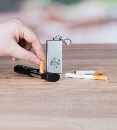 Bedruckter Taschenascher in Metall stehend und Taschenaschenbecher in Kunststoff liegend auf einem Tisch, daneben zwei Zigaretten und eine Hand, die die Zigarette im personalisierten Taschenascher ausdämpft