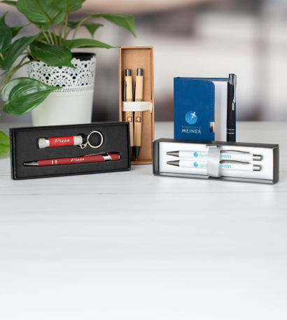 Schreibsets mit individuellem Branding liegen und stehen auf einer weißen Oberfläche neben einer Grünpflanze in einem Topf
