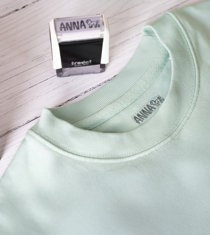 Textilstempel mit individuellem Motiv steht auf einem Tisch und daneben liegt ein türkises T-Shirt mit dem darauf gestempelten Namen 