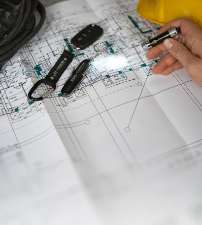 Verschiedene Schlüsselanhänger mit personalisiertem Logo liegen auf einem CAD-Plan und eine Person hat einen Schlüsselanhänger mit Taschenlampenfunktion in der Hand und leuchtet auf den Plan