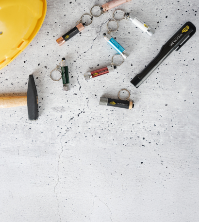 Bedruckte Taschenlampen als Schlüsselanhänger und Taschenlampen mit Clip liegen auf Beton neben einem Hammer und einem gelben Helm