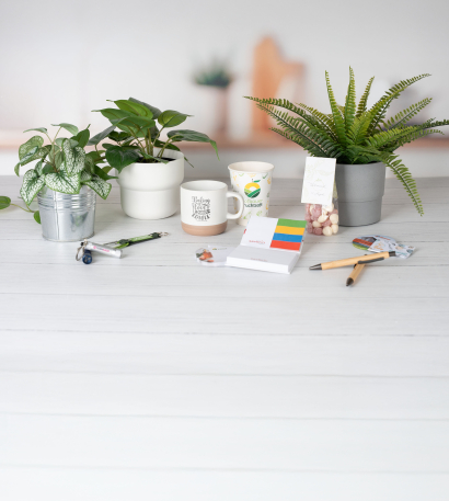 Verschiedene bedruckte und personalisierte Werbeartikel stehen neben Grünpflanzen im Topf und auf weißem Untergrund