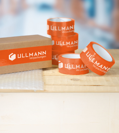 Paketklebebänder auf Rollen im individuellen Design stehen aufgetürmt neben einem Paket, das mit einem selbstgestalteten Paketklebeband zugeklebt wurde auf einem Bürotisch