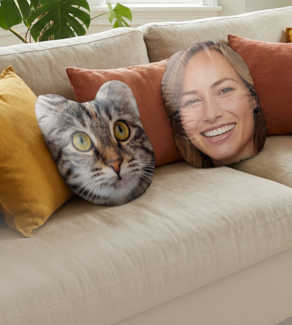 Ein Gesichtspolster einer weiblichen Person und ein Gesichtskissen in Form einer Katze liegen auf einem beigen Sofa