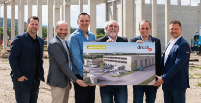 Geschäftsführung von druck.at steht vor Baustelle mit einer Tafel in den Händen, worauf der 3D-Plan des neuen Firmengebäudes darauf zu sehen ist.