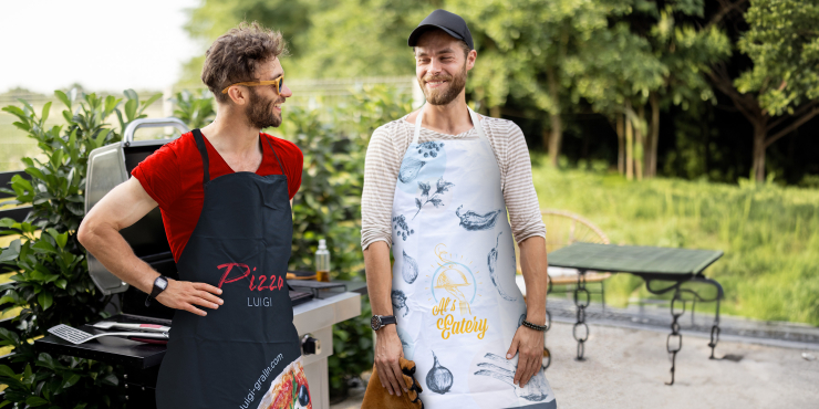 zwei Männer mit verschiedenen individuell bedruckter Kochschürze neben Grill