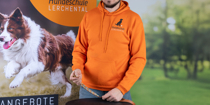 Hundeschulen-Trainer trägt einen orangenen B&amp;C™ Hoodie mit Bauchtasche und aufgedrucktem Logo auf der linken Brustseite