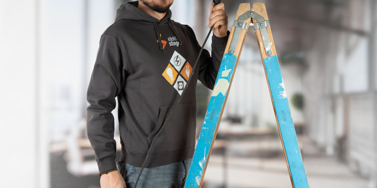 Elektriker steht neben Leiter und trägt einen Hoodie mit Bauchtasche in der Farbe Anthrazit mit dem aufgedruckten Firmenlogo auf der Vorderseite
