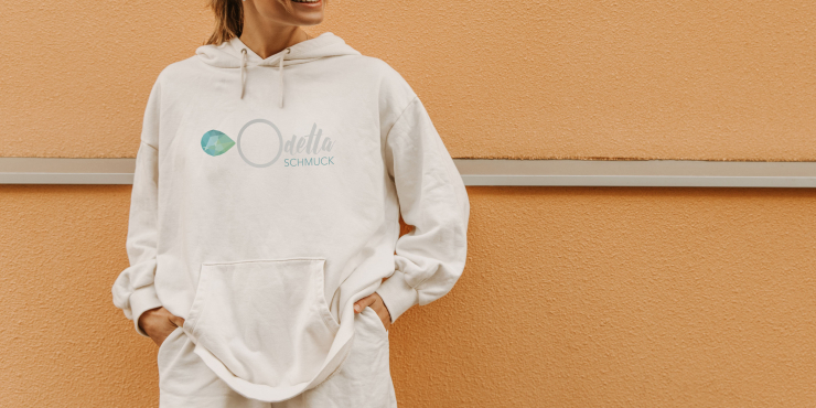 Frau steht vor Hausmauer und trägt einen weißen Hoodie mit einem Logo auf der Vorderseite aufgedruckt