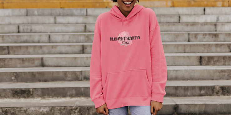 Weibliche Person steht vor einer Treppe und trägt einen rosafarbenen Fruit of the Loom-Hoodie mit Logo auf der Vorderseite aufgedruckt 