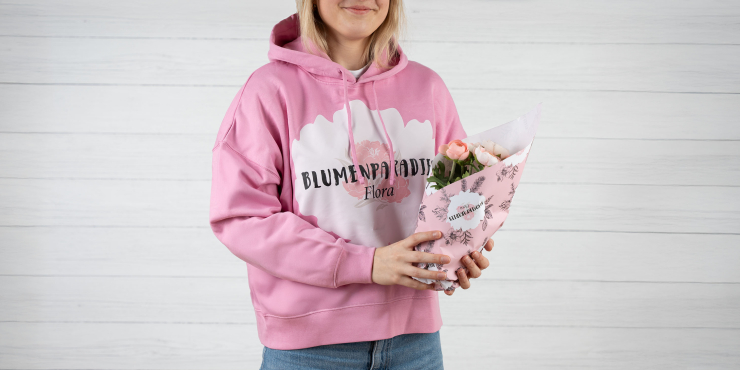 Mädchen mit rosarotem Hoodie, der mit einem Logo personalisiert wurde, steht vor grauem Hintergrund und hält einen Blumenstock