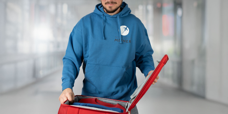 Mann steht vor einem geöffneten Koffer und trägt einen blauen Stanley/Stella-Hoodie mit Aufdruck auf der linken Brustseite