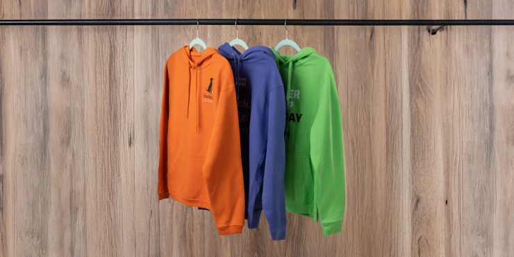 Orangener, dunkellila und grasgrüner Hoodie bedruckt mit Motiven hängen auf einer Stange