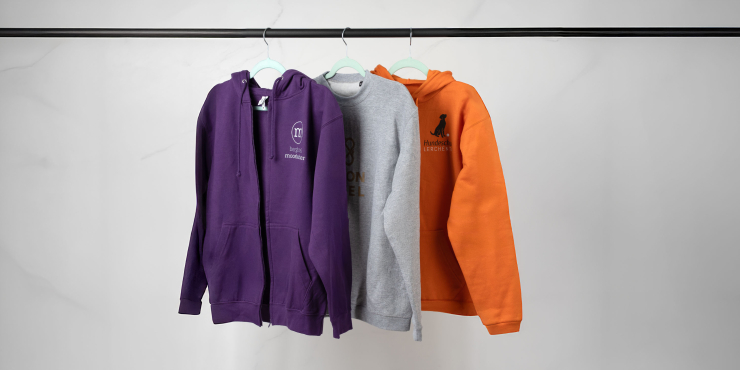 Dunkellilafarbener Zip-Hoodie, grauer Sweater und orangener Hoodie sind mit verschiedenen Designs bedruckt und hängen auf einer Stange