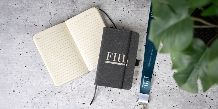 Carson A6-Notizbuch personalisiert liegt geschlossen und offen auf einem Betonboden neben einer Grünpflanze und einem schwarzen Lanyard