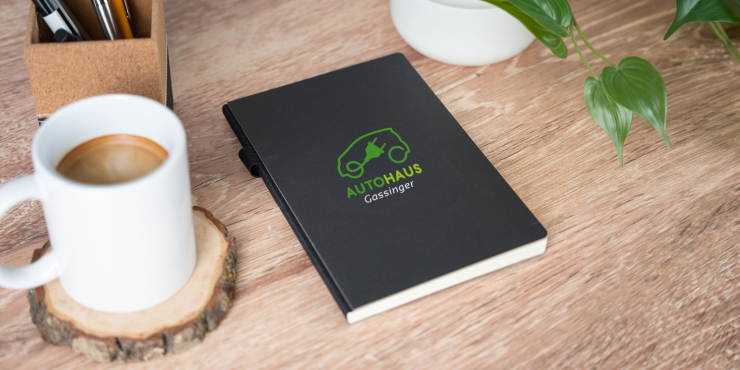 Schwarzes Cover Journal Notizbuch im A5-Format und individuellem Design liegt neben einer Grünpflanze und einer Kaffeetasse auf einem Bürotisch 