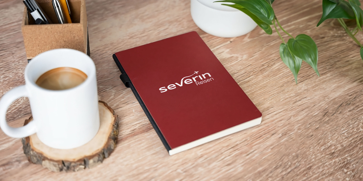 Rotes Cover Journal Notizbuch im A5-Format und individuellem Design liegt neben einer Grünpflanze und einer Kaffeetasse auf einem Bürotisch 