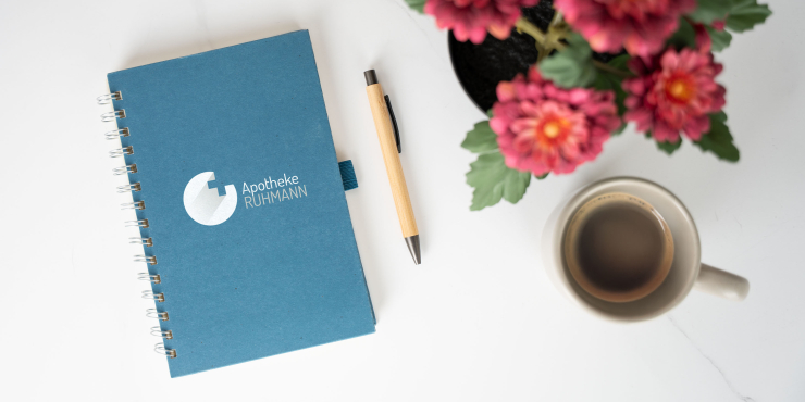 Terra Notizbuch mit Spiralbindung und personalisiertem Design liegt geschlossen neben einem Kugelschreiber und einer Tasse Kaffee und einem Blumentopf mit roten Blumen