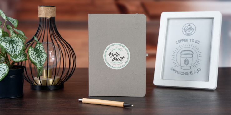 Terra Notizbuch im A5-Format und im personalisiertem Design steht neben einem Bilderrahmen, neben einer Lampe und einer Pflanze auf einem Tisch