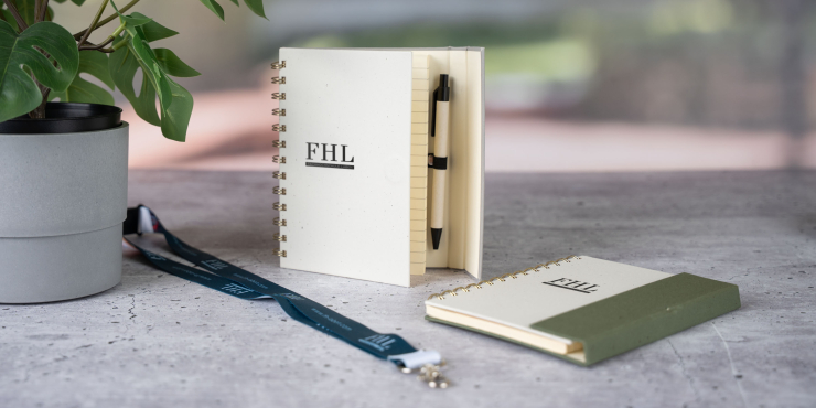 Halden-Notizbuch mit Kugelschreiber und Haftnotizen steht neben einer Grünpflanze auf einem Tisch, daneben liegt ein Lanyard