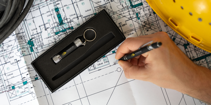 Person zeichnet mit personalisiertem Kugelschreiber auf einem Plan, daneben liegt die geöffnete Schachtel, worin eine Taschenlampe als Schlüsselanhänger liegt