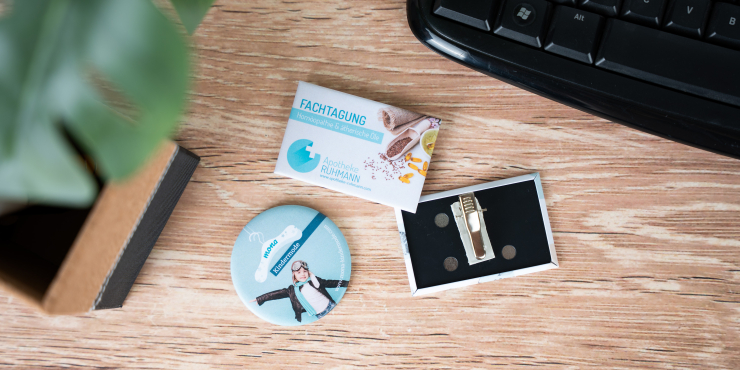 Runder und rechteckiger Button mit Clip individuell gestaltet liegen auf einem Schreibtisch neben einer schwarzen Tastatur und einer Grünpflanze