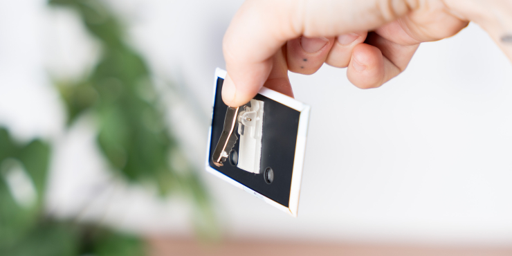 Person zeigt die Rückseite des Clip-Buttons und öffnet den Clip mit dem Daumen, im Hintergrund steht eine Grünpflanze