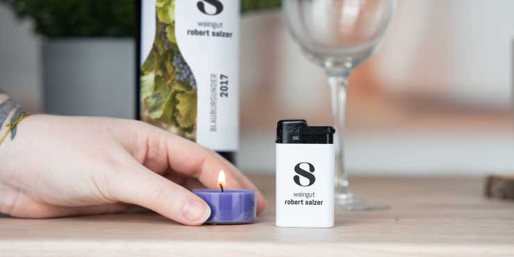 Person stellt eine brennende Kerze neben das GO Elvo Feuerzeug vor eine Flasche Wein