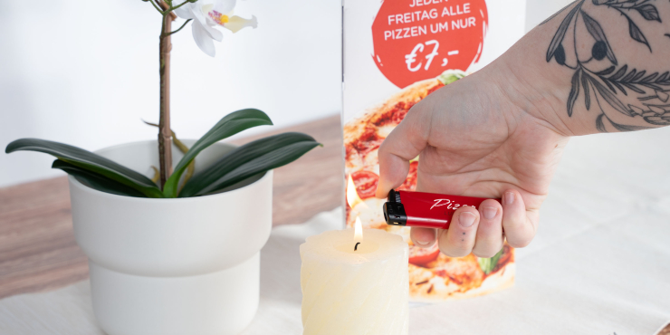 Person zündet mit bedrucktem Feuerzeug eine Kerze an, die neben einer Pflanze steht