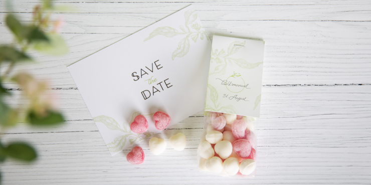 Herzbonbons in individuell gestalteter Verpackung liegen auf weißem Holzuntergrund und Einladungskarte