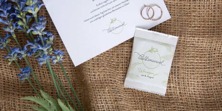 Personalisierte Verpackung gefüllt mit Kaugummi auf bräunlichem Untergrund liegend neben Flyer und Blumen