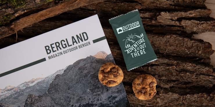 Zwei Schokoladenkekse neben einer individuell gestalteten Verpackung und einem Outdoor-Magazin, auf einer Holzoberfläche