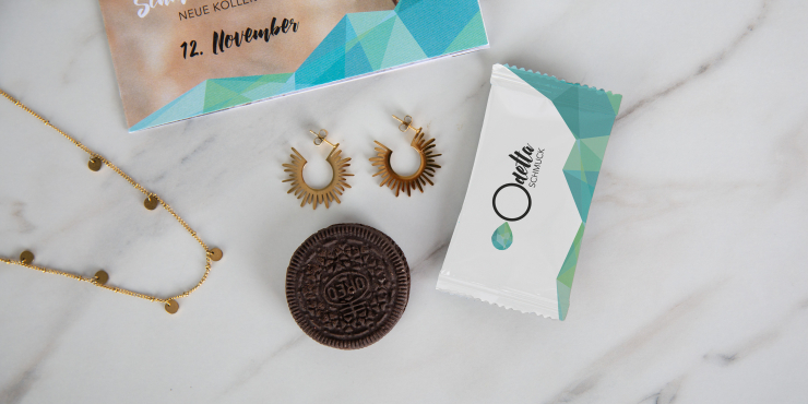 Ein Schokoladenkeks neben einer individuell gestalteten Verpackung, Schmuckstücken und einem Schmuckkatalog auf einem Marmortisch