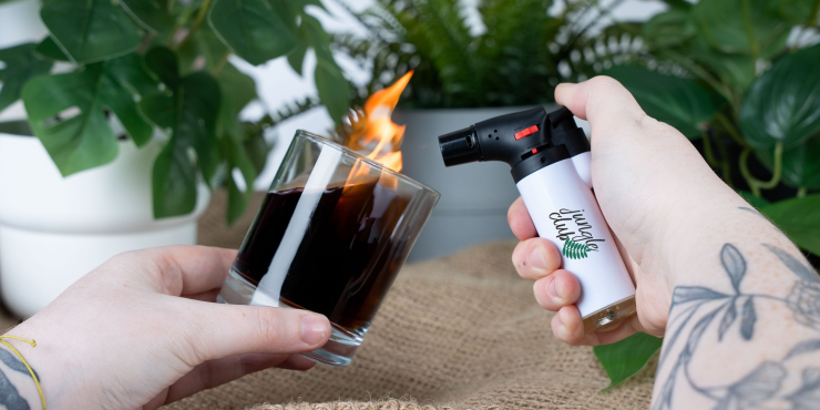Person hält ein Glas und das bedruckte LUX Jetflame Feuerzeug in der Hand vor Blumen