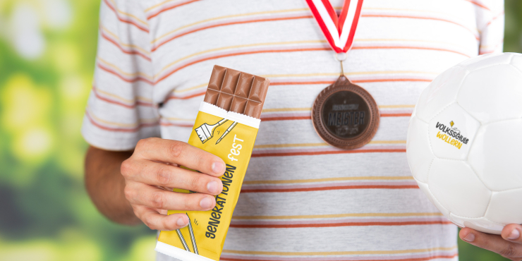 Person in gestreiftem T-Shirt hält eine individuell gestaltete Schokolade und einen Fußball, trägt eine Medaille