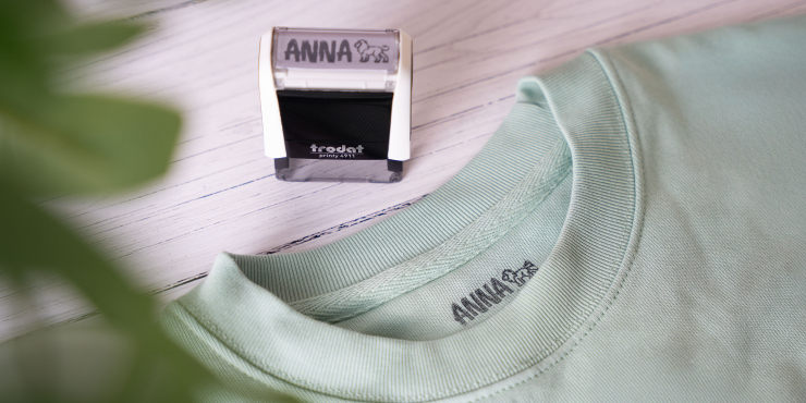 Trodat Printy Textilstempel mit eigenem Logo steht auf einem Tisch neben einem mintgrünem T-Shirt, worauf der Name Anna aufgestempelt ist