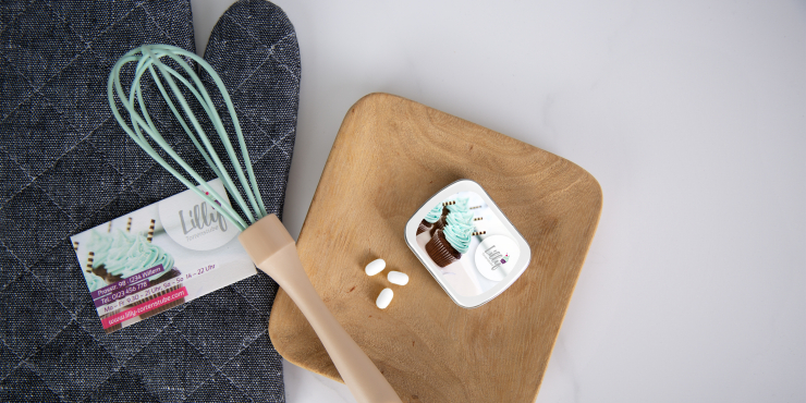 Backhandschuh, Schneebesen und Visitenkarte liegen neben einem hölzernen, quadratischen Teller, worauf eine personalisierte Klappdose liegt, die mit TicTacs gefüllt ist 