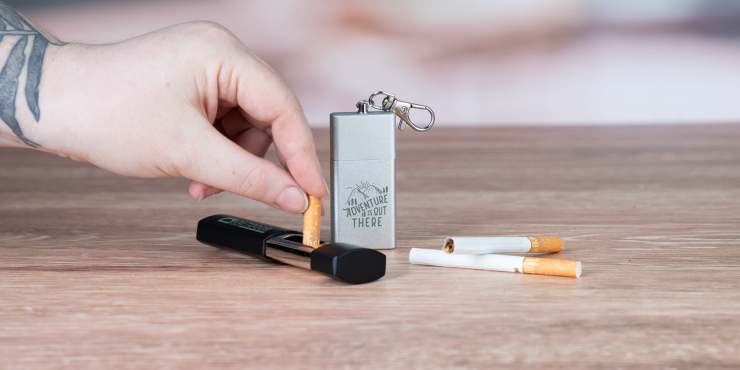 Taschenaschenbecher aus Kunststoff liegend auf einem Tisch neben bedruckten Taschenaschenbecher aus Metall, wo jemand seine Zigarette ausdämpft