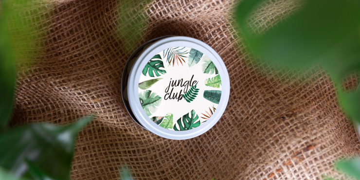 Runde Dose mit &#039;jungle club&#039; Design und Blättermuster auf Jutestoff, umgeben von grünen Blättern