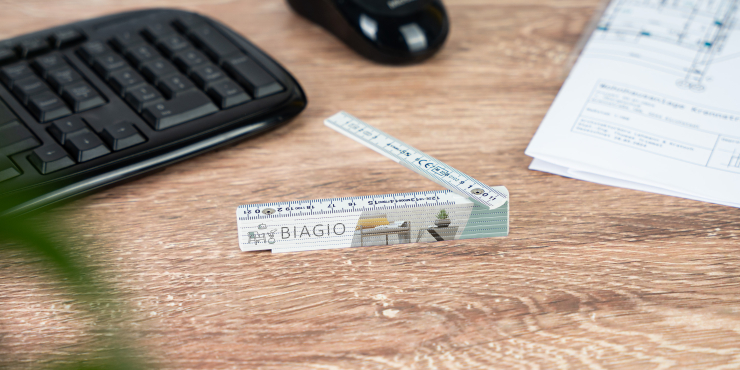 Personalisierter Ein-Meter-Zollstock liegt auf Bürotisch neben Tastatur, Notizblock und Computermaus