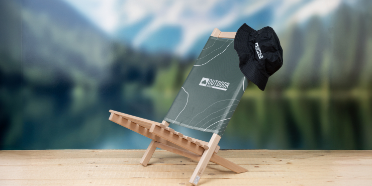 Klapp-Liegestuhl bedruckt mit personalisiertem Design stehend auf einem Steg mit schwarzem Fischerhut, im Hintergrund liegt ein See am Fuße eines Berges