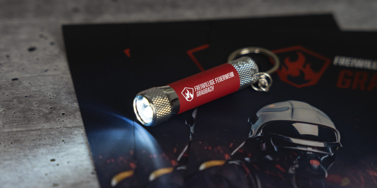 Eine bedruckte Taschenlampe mit Schlüsselring liegt auf einem Flyer