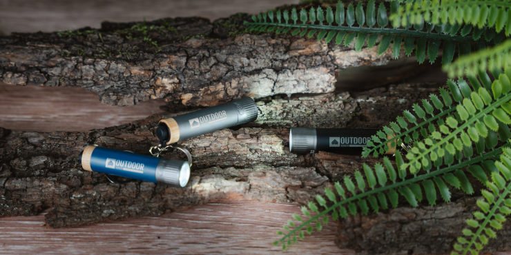 Mehrere bedruckte Taschenlampen mit Schlüsselring liegen in einem Wald